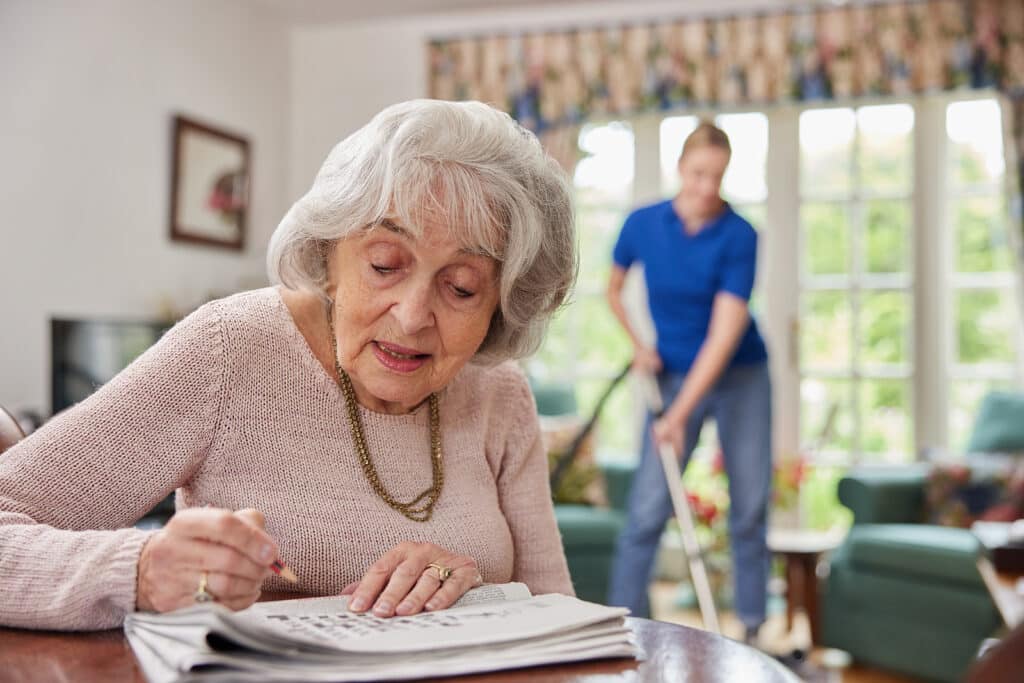Older woman in a pink sweater reads a newspaper at a table; a man vacuums in the sunny living room behind her.